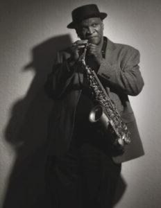 A black and white picture of an African American man posing in a black hat, holding a saxophone